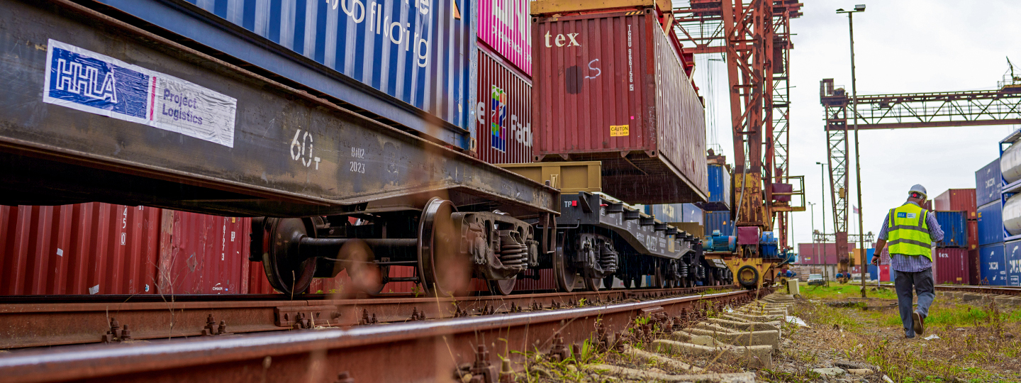Güterzug beladen mit Containern, im Hintergrund ein Kran und ein HHLA-Mitarbeiter, der weggeht (Foto)